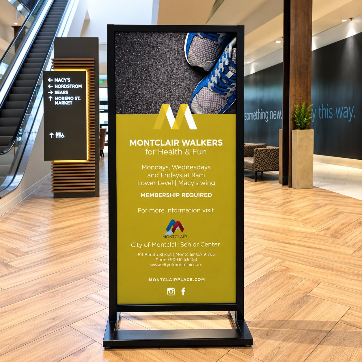 A free-standing Montclair Walkers wayfinding sign in a shopping mall, with a yellow panel listing event details and an upper panel showing a pair of sneakers on asphalt.