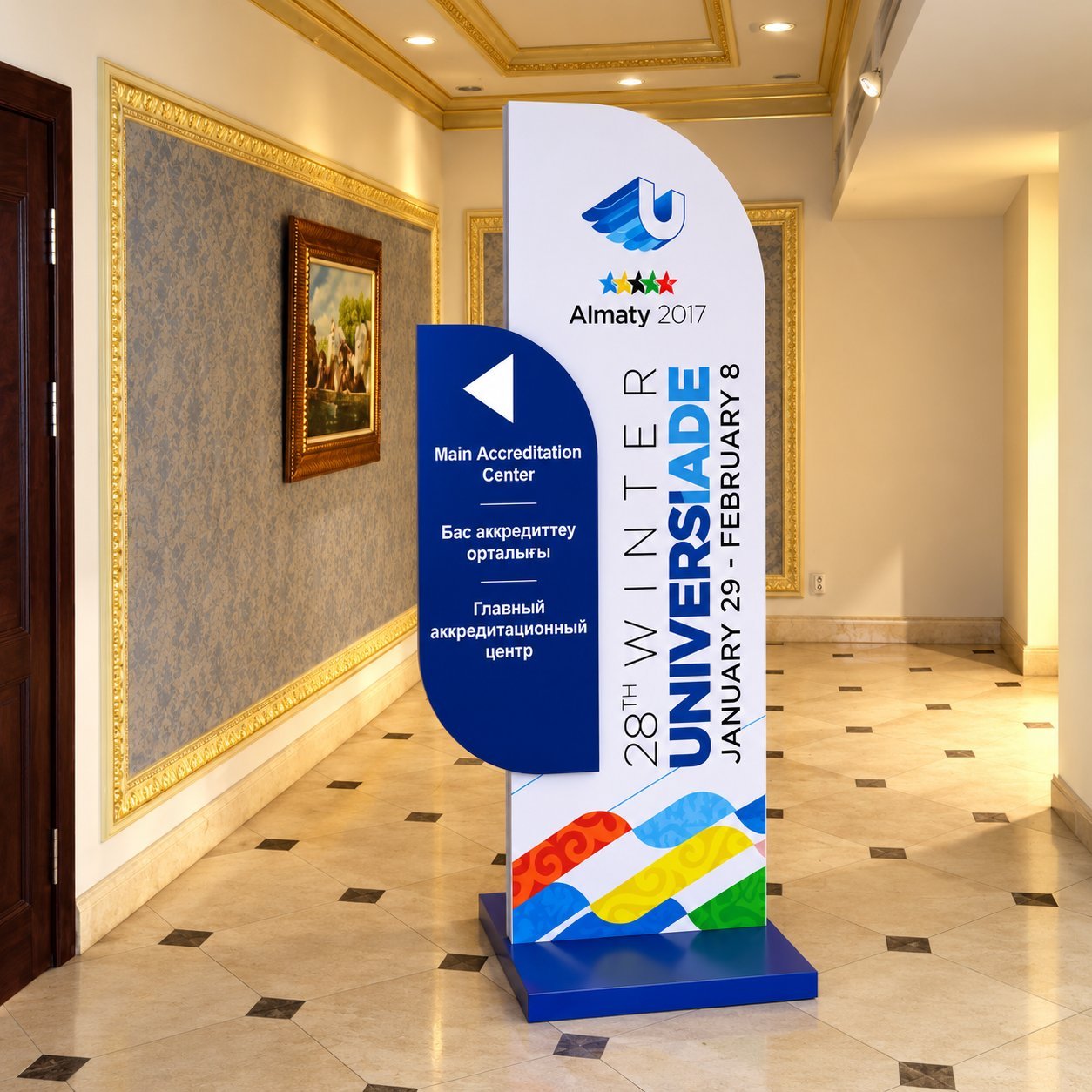 A blue-and-white 28th Winter Universiade Almaty 2017 event directional sign in a corridor, with the Main Accreditation Center arrow and multilingual text.
