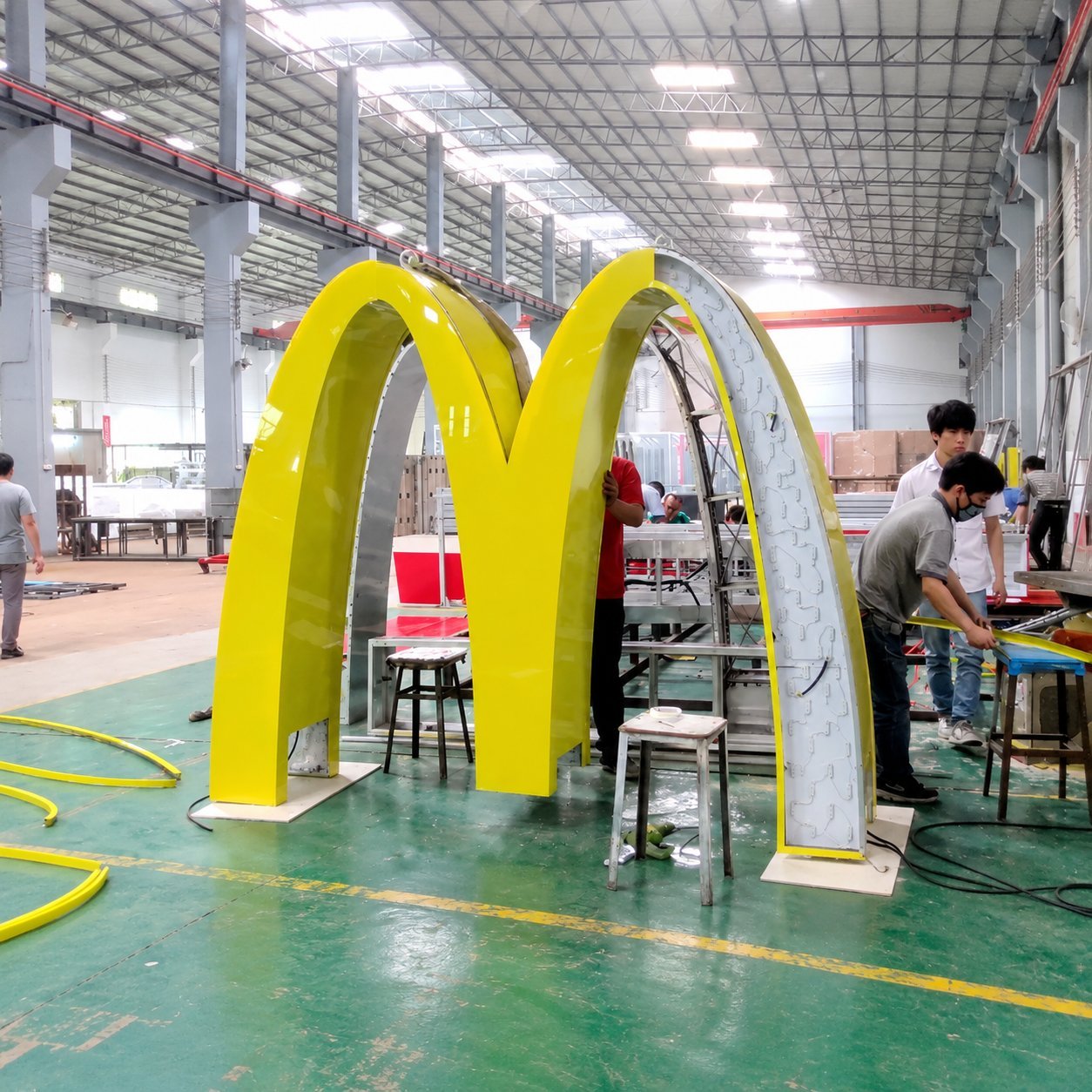Large yellow illuminated arch sign under fabrication in a Hicen factory, with workers assembling the structure on the shop floor.