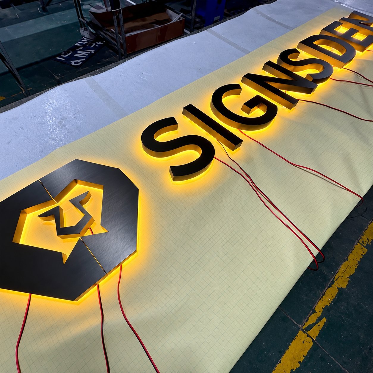 Illuminated SIGNED channel letters with house-shaped logo under final bench test, glowing warm white with red safety-marking wiring visible.