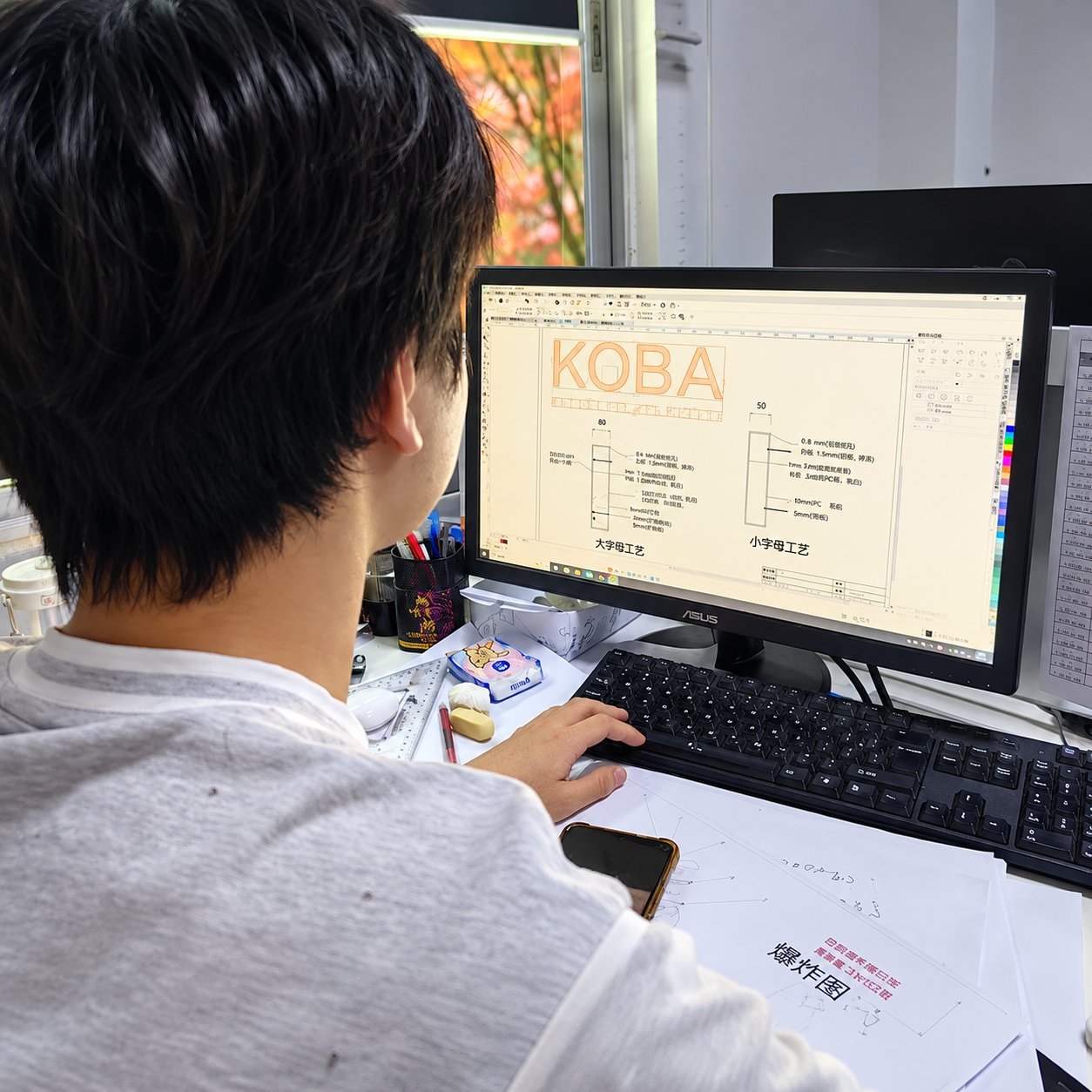 Sign designer working at a monitor, refining a vector drawing of a 'KOBA' logo in CAD with annotations for letter height and material thickness.